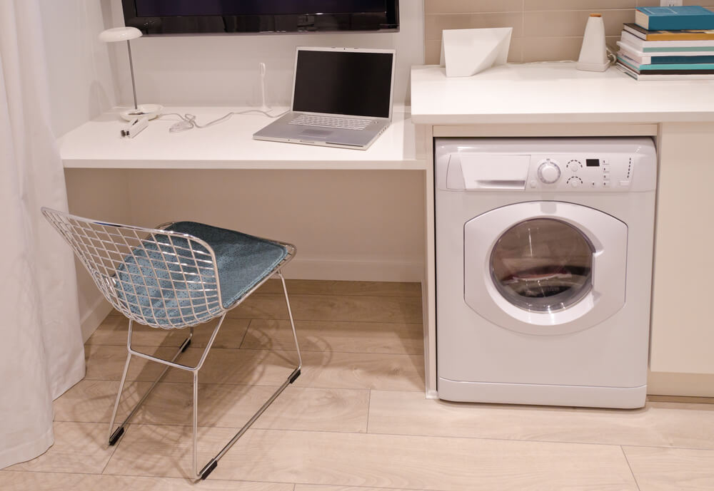 Foto que ilustra matéria sobre lavanderia pequena planejada mostra uma máquina de lavar roupas abaixo de uma bancada em um home office. Ao lado do aparelho, uma cadeira. E na continuação da bancada há um notebook.