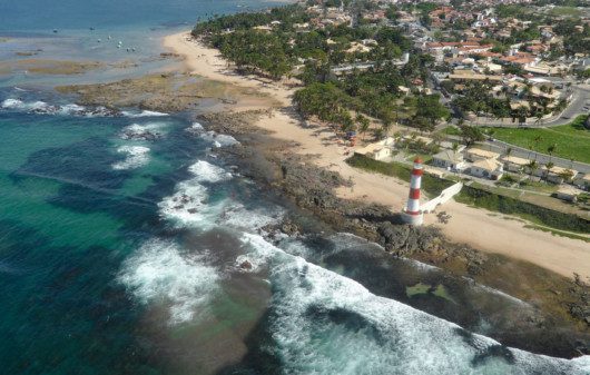 Foto que ilustra matéria sobre as praias de Salvador mostra uma vista aérea da Praia de Itapuã, com o mar ocupando parte da tela do meio para a esquerda, o famoso farol de Itapuã no meio e a faixa de terra mais para o canto superior direito da imagem.