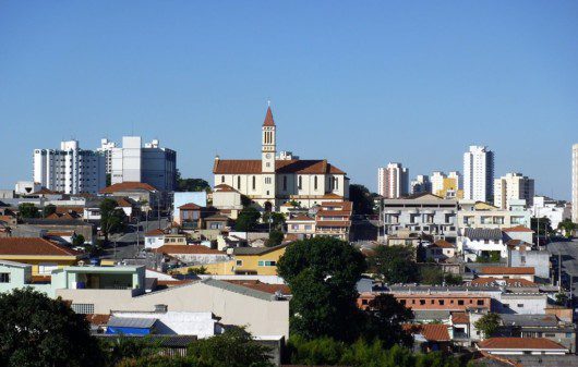 Foto que ilustra matéria sobre a Zona Leste de SP mostra uma panorâmica do bairro de Vila Matilde, com uma igreja ao fundo, no centro da tela, e um céu azul.