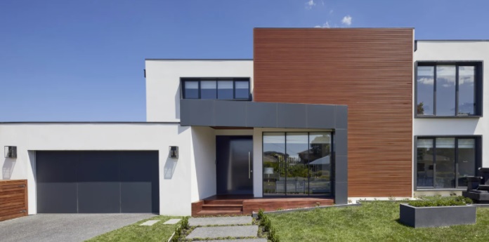 A imagem mostra uma casa quadrada de dois andares com janelas de vidros e portão da garagem cinza escuro. A madeira é utilizada em um dos elementos retangulares, reforçando listras geométricas.