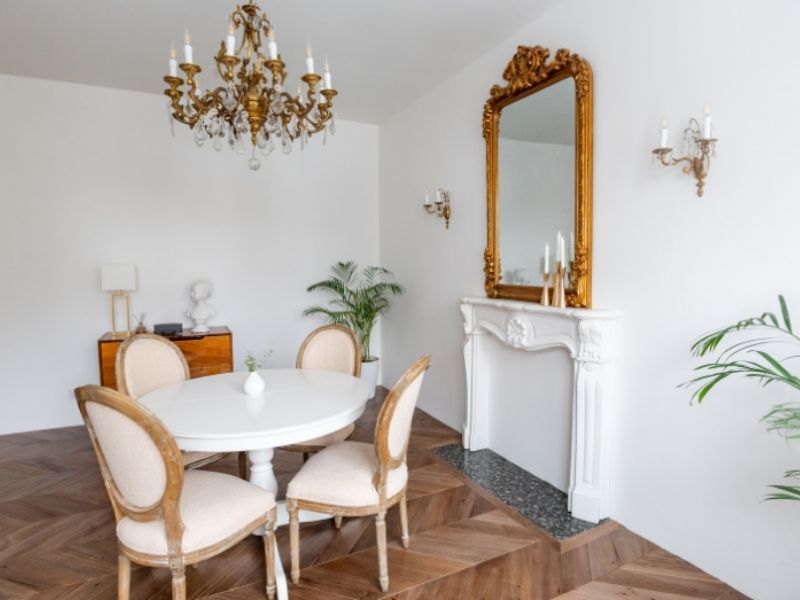 Sala com mesa de 4 lugares e um candelabro elaborado. Na parede, um espelho com moldura dourada ornamentada.