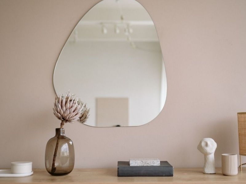 Espelho minimalista, na parede. Abaixo dele há um suporte com um vaso de flores e itens decorativos.