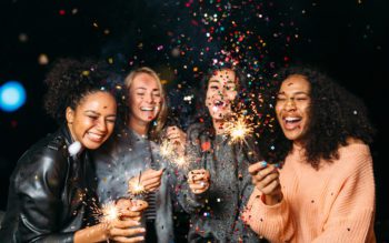 Foto que ilustra matéria sobre resoluções de ano novo mostra quatro jovens mulheres em uma festa de Réveillon, sorridentes e com velas que soltam faíscas nas mãos, enquanto confetes coloridos voam pelos ares diante delas.