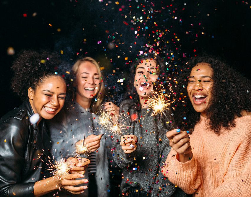 Foto que ilustra matéria sobre resoluções de ano novo mostra quatro jovens mulheres em uma festa de Réveillon, sorridentes e com velas que soltam faíscas nas mãos, enquanto confetes coloridos voam pelos ares diante delas.