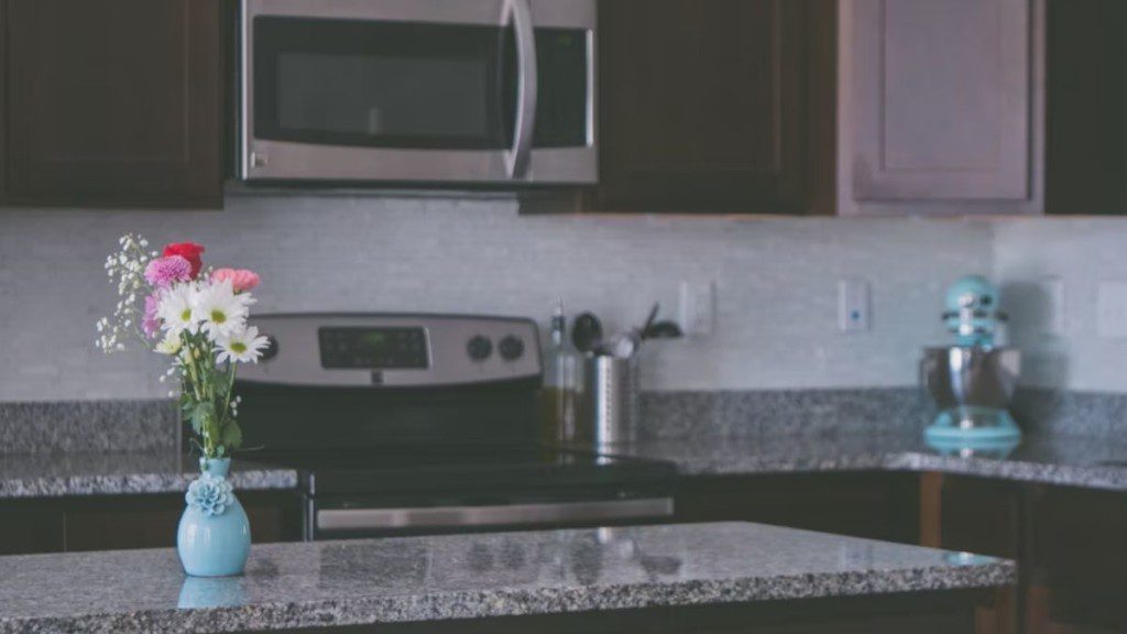 Imagem de um balcão de cozinha americana de granito, com um jarro azul de flores acima. Ao fundo, um fogão e, em cima dele, um microondas.