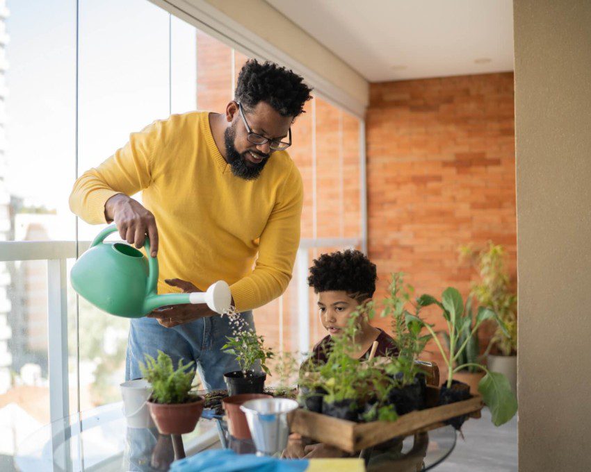 Foto que ilustra matéria sobre Como fazer horta em casa mostra um homem negro, de camisa amarela comprida, usando um regador para molhar pequenos vasos de plantas com ervas ao lado de seu filho em uma sacada de apartamento