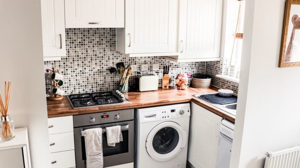 A foto mostra uma cozinha pequena com parede texturizada, móveis (bancada, armários e gavetas) e utensílios (fogão, pia, máquina de lavar, torradeira, faqueiro, canecas e outros). 