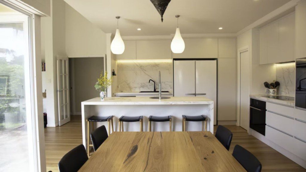 A foto mostra uma cozinha americana pequena com mesa no centro e bancada dividindo ambientes. Na imagem há também dois lustres de teto, uma pia ao fundo, geladeira de duas portas, banquetas e cadeiras, entre outros elementos.