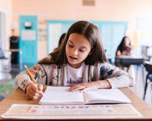 Foto que ilustra matéria sobre Escolas em Vitória (ES) mostra uma menina sentada com um caderno aberto à sua frente e escrevendo com um lápis amarelo em uma sala de aula.