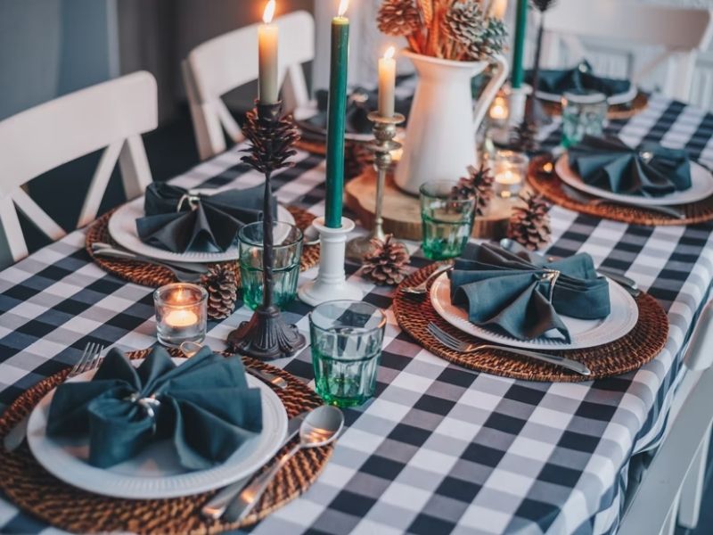 mesa posta com toalha xadrez, pratos acima de jogo americano marrom, talheres aos lados dos pratos, guardanapos azuis acima. Há velas e uma jarra.