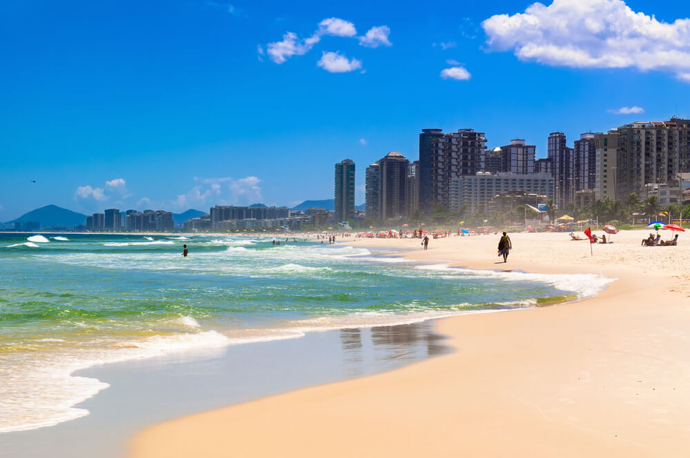 Foto que ilustra matéria sobre as praias do Rio de Janeiro mostra uma visão da faixa de areia da praia da Barra da Tijuca, com a água do mar à esquerda e prédios à direita e mais ao fundo da imagem.