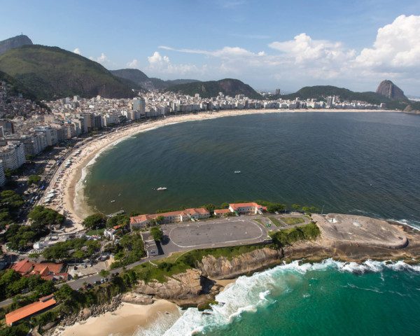 Foto que ilustra matéria sobre as praias do Rio de Janeiro mostra uma visão aérea de toda a orla da praia de Copacabana, a começar pelo Forte de Copacabana, em primeiro plano, mais abaixo na tela, seguido de toda a faixa de areia e boa parte do mar, com os prédios à esquerda e montanhas ao fundo. Entre elas os morros do Corcovado e do Pão de Açúcar