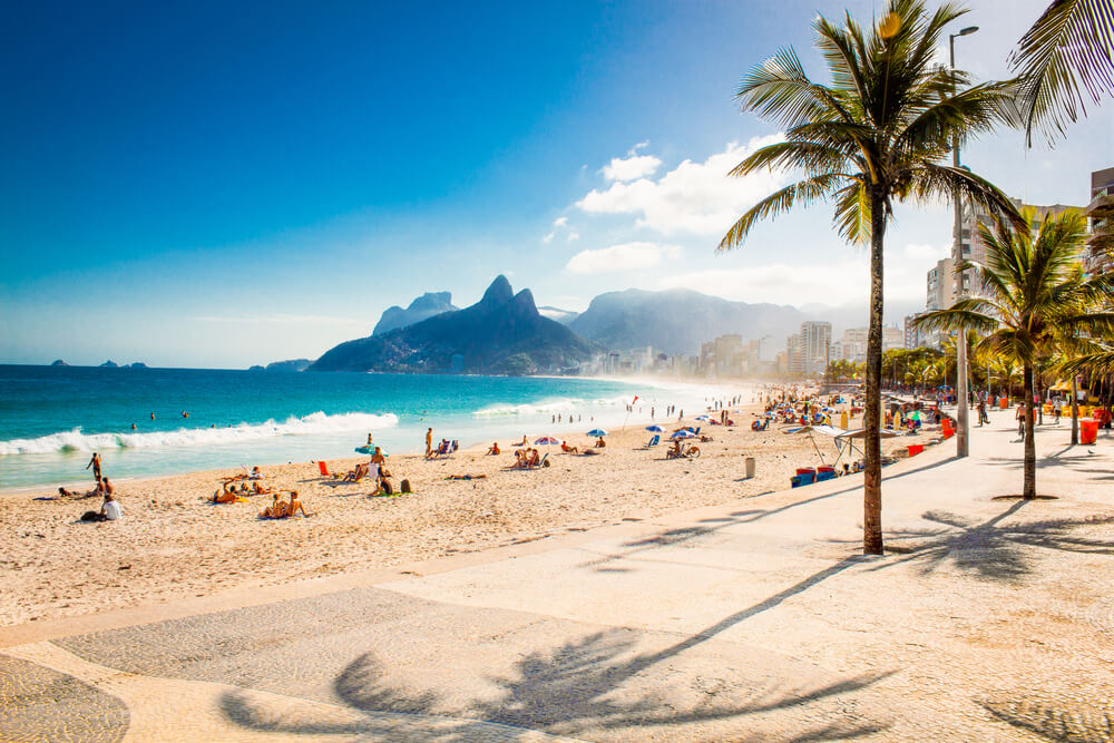 Foto que ilustra matéria sobre as praias do Rio de Janeiro mostra uma visão tirada da calçada da praia de Ipanema, com a faixa de areia ao centro, o mar na sequência e o morro Dois Irmãos ao fundo.