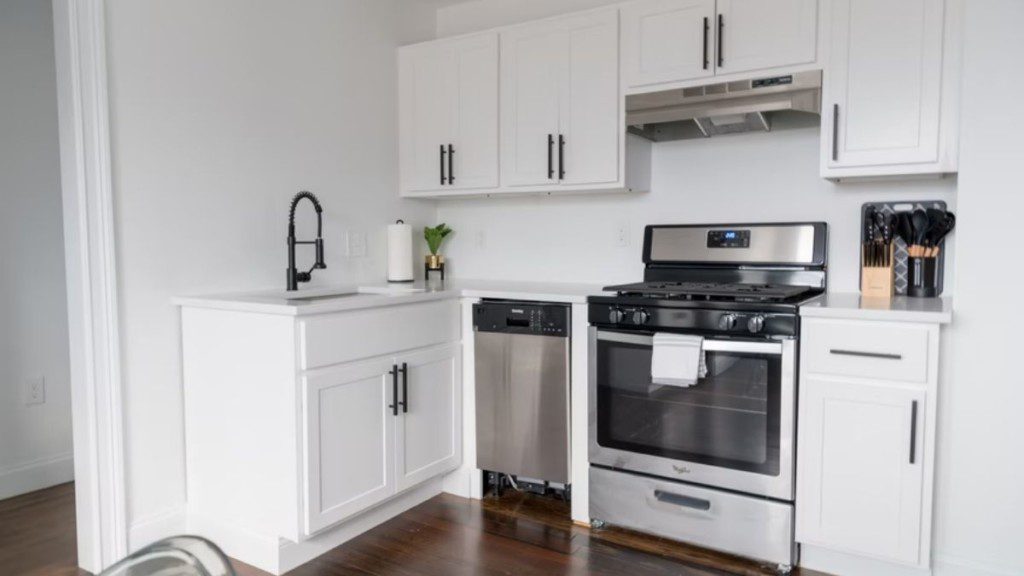 A foto mostra uma cozinha pequena em tons brancos e neutros. Na imagem há: armários, fogão com coifa, pia e alguns utensílios como faqueiro e colheres.