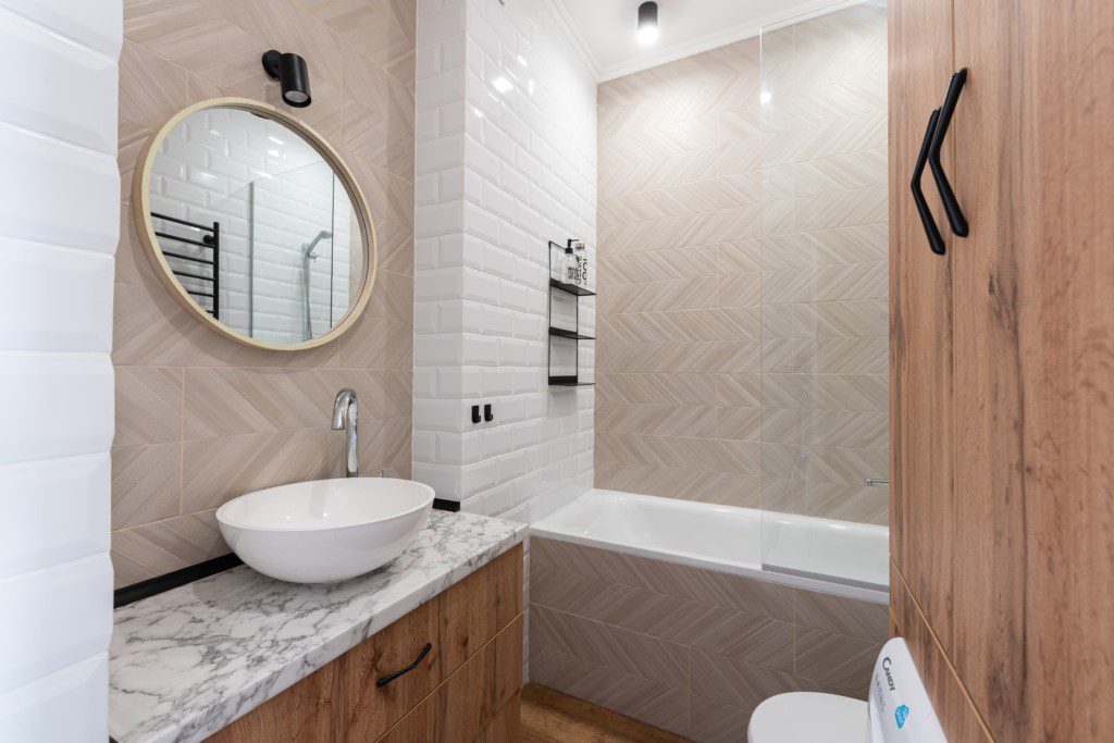 Foto mostra um banheiro pequeno com uma banheira branca. Há também um espelho oval, uma pia e armários embutidos nela.