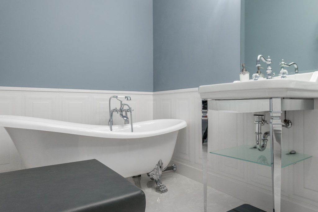 Foto de um banheiro pequeno com banheira minimalista. Na imagem há uma banheira retangular com bordas arredondadas. Dentro dela há uma torneira com ducha. Há também uma pia com torneira na imagem.