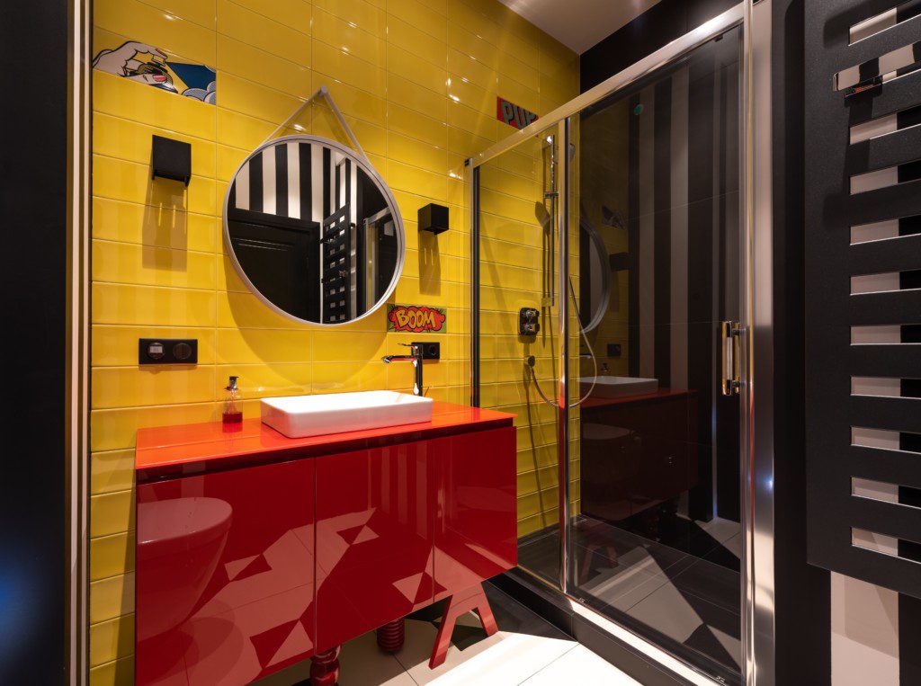A foto mostra um banheiro pequeno decorado com cores fortes: parede amarela, armário vermelho, área para banho em preto. Há também um espelho oval e uma pia branca.
