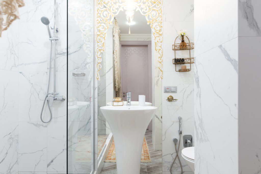 Foto de um banheiro pequeno com detalhes dourados no espelho. Há também uma pia, box de vidro transparente, ducha e vaso sanitário.