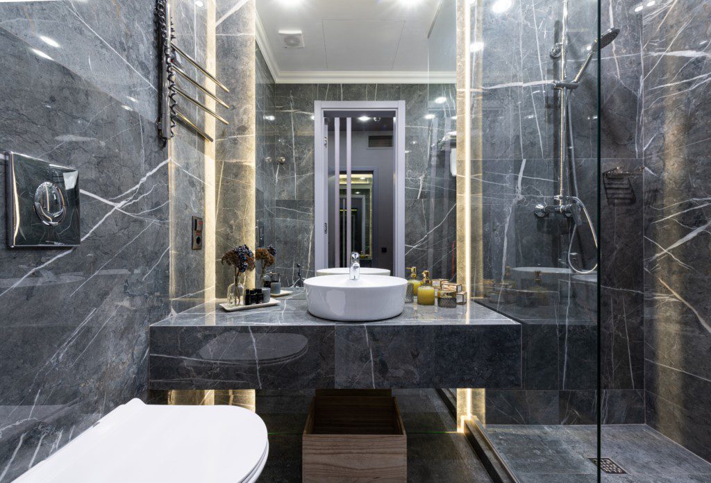 A foto mostra um banheiro pequeno todo em tons de preto com detalhes dourados feitos pela iluminação. Há na imagem uma bancada preta com uma pia branca em cima, um espelho grande, um vaso sanitário e uma área para banho.