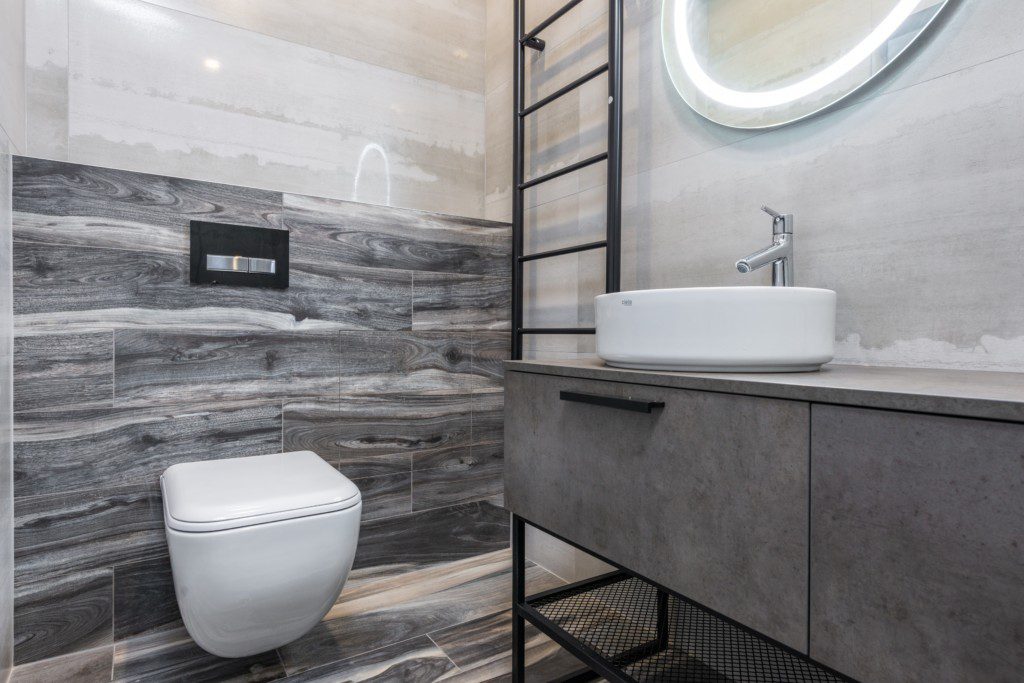 Foto de um banheiro pequeno decorado com estilo industrial. Nele há: vaso sanitário, bancada cinza, pia, espelho oval e suporte para toalhas.