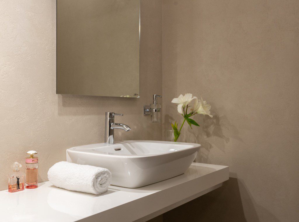 Foto de um banheiro pequeno decorado com uma flor. Nele há: espelho retangular, pia, itens de higiene pessoal e duas rosas brancas.