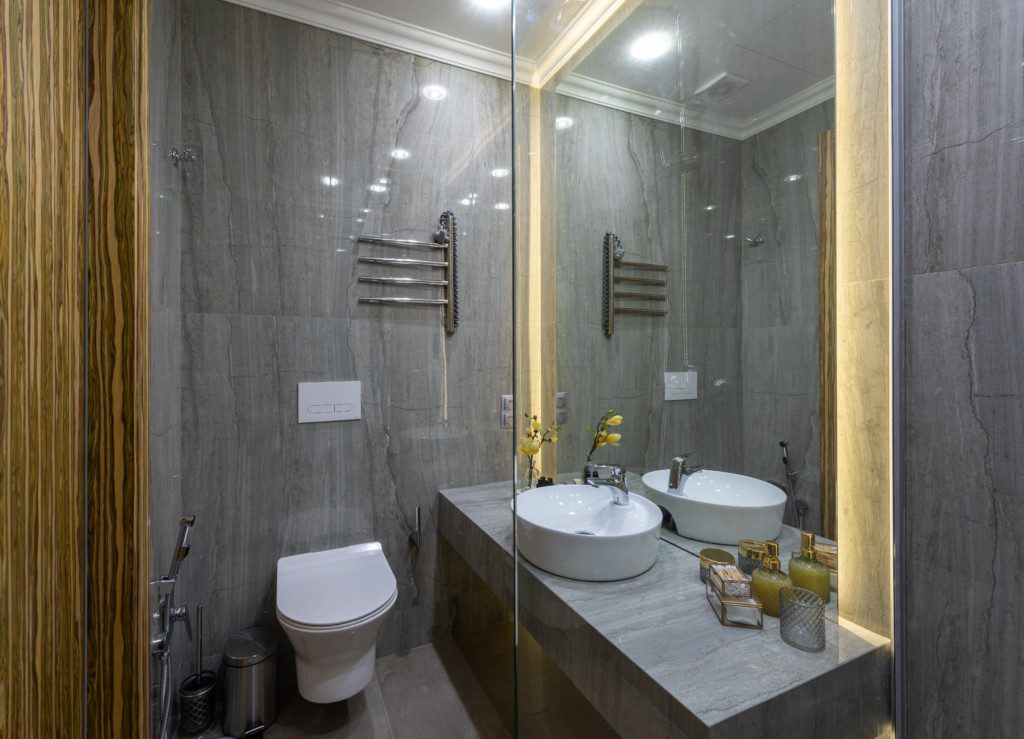 A foto mostra um banheiro com iluminação especial nas laterais do espelho principal. Há também um vaso sanitário, uma pia e suporte para pendurar toalhas.