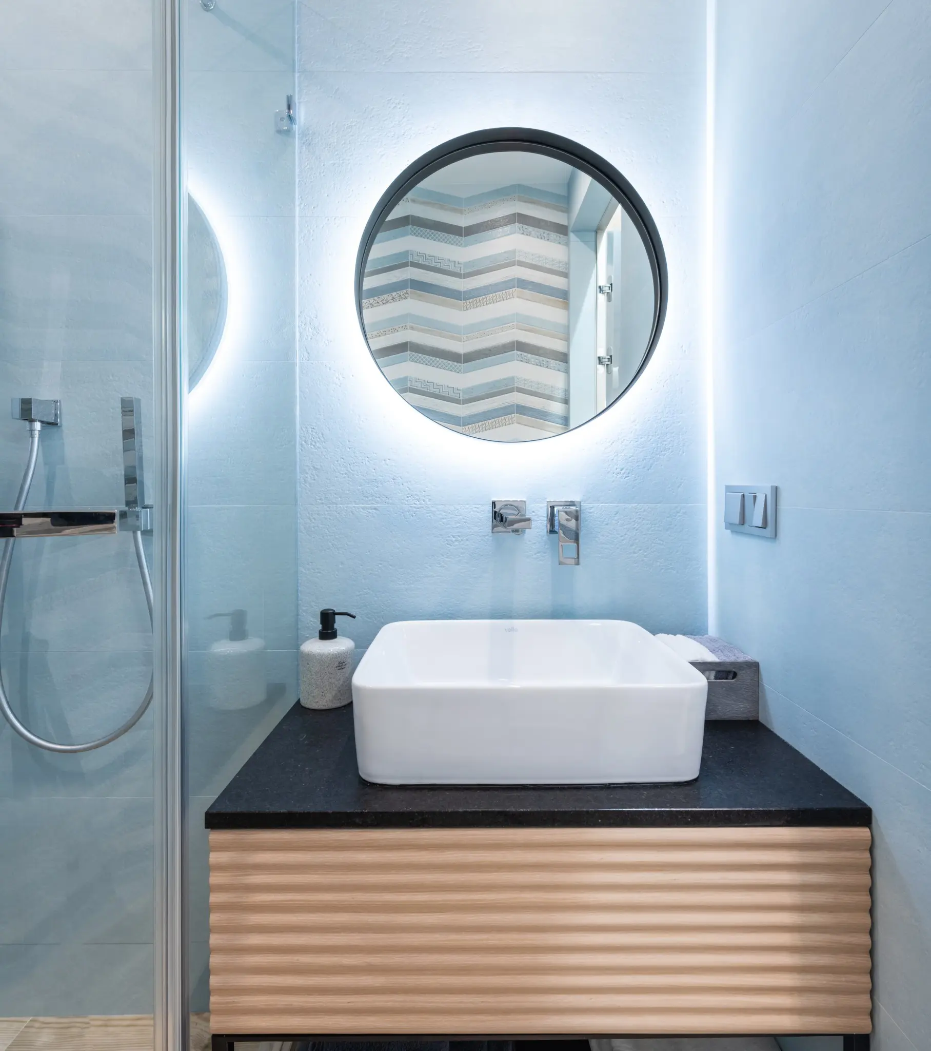 A foto mostra um banheiro pequeno bem iluminado e decorado com fita de led ao redor do espelho redondo.
