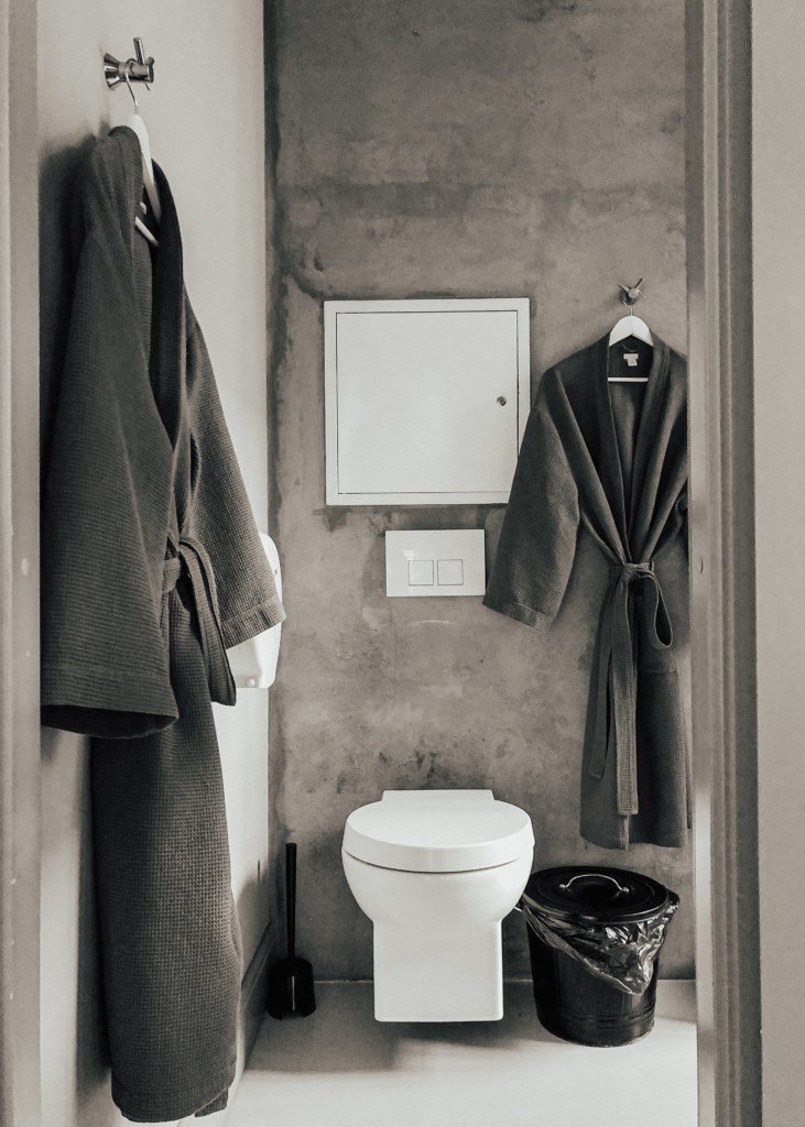 Foto de banheiro pequeno que usa dois registros de água como suporte para pendurar dois roupões de banho (um em cada registro). Há também na imagem um vaso sanitário e lixeira.