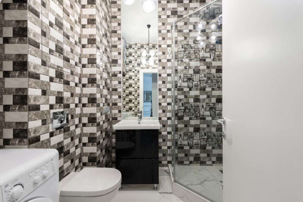 Foto de um banheiro pequeno decorado com textura. A textura está na parede, com tijolinhos de diferentes cores: banco, cinza e preto. Há também na imagem vaso sanitário, pia, armário, espelho e área para banho.