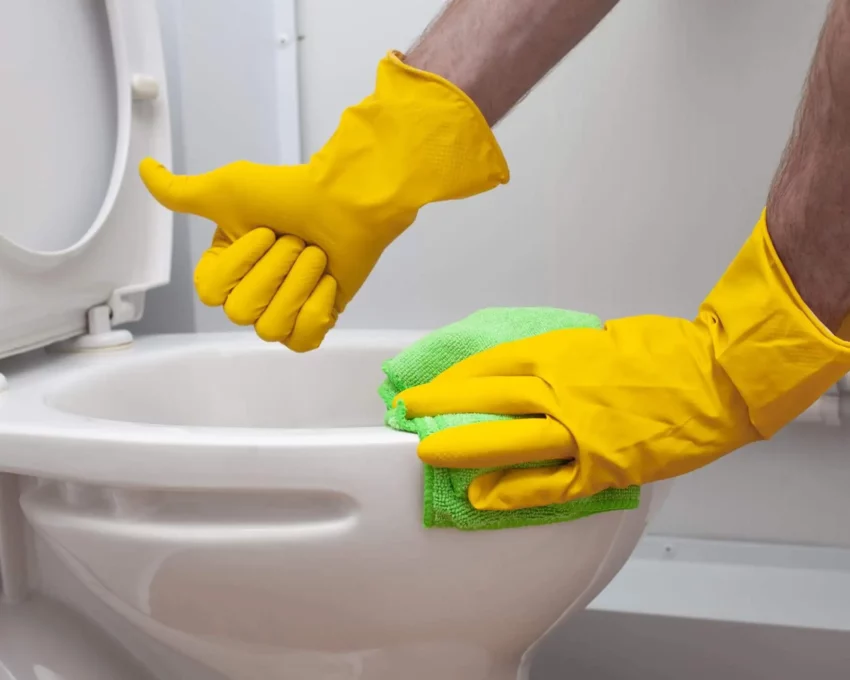 Imagem de um homem limpando um vaso sanitário com luvas amarelas e fazendo sinal de joinha para ilustrar matéria sobre como desentupir vaso sanitário