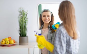 Foto que ilustra matéria com dicas sobre como limpar espelho mostram uma mulher de costas, posicionada de frente para um espelho. Ela aparece sorrindo no reflexo, com um luvas amarelas, com um pano verde esfregando o espelho em uma das mãos e um borrifador na outra.