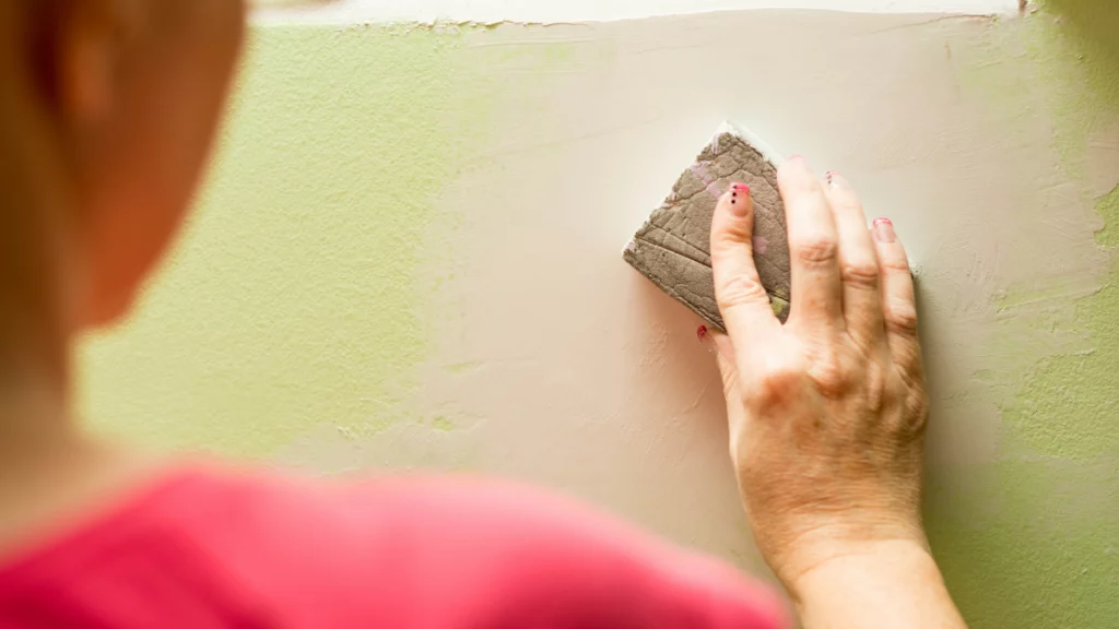 Foto de uma pessoa de costas. A pessoa está lixando uma parede verde.