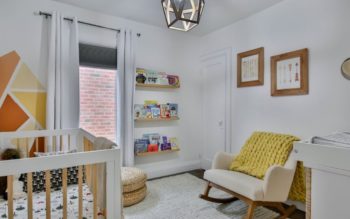 quarto de bebê decorado em tons de branco e mostarda. Há um berço, uma janela coberta por cortinas, uma poltrona e uma cômoda.