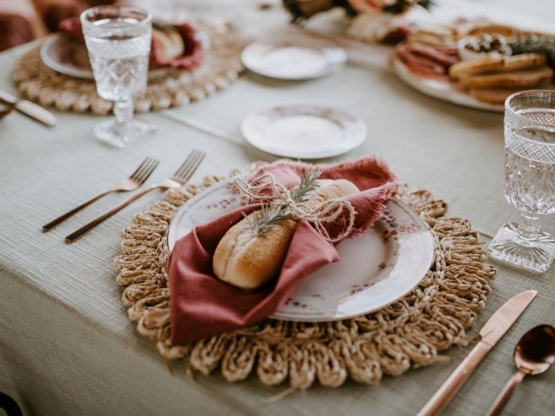 mesa posta com jogo americano circular, guardanapo rosado sobre os pratos, talheres de sopa, entrada, sobremesa e prato principal. Pratos de sobremesa ao centro.
