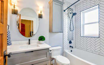 Banheiro pequeno minimalista em tons de branco, preto e cinza com espelho redondo, banheira, luminárias de parede e móveis planejados.