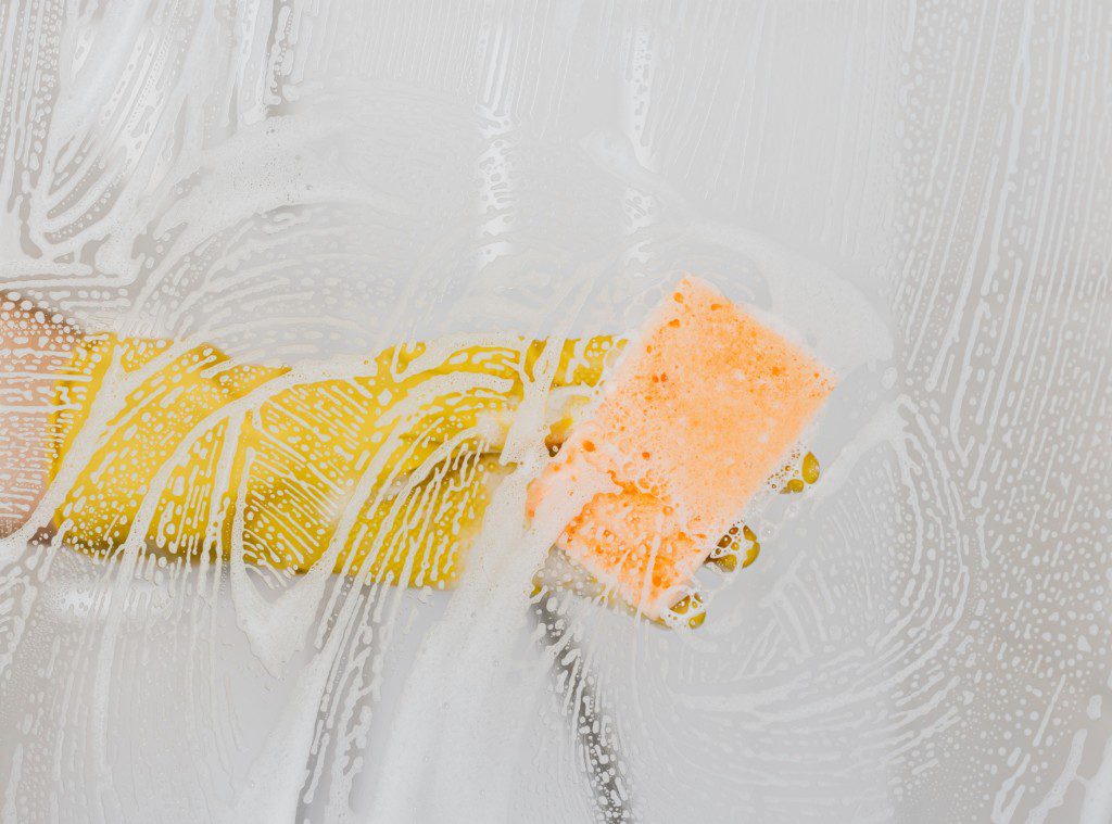 Foto que ilustra matéria sobre como limpar box mostra uma mão com luva amarela passando uma esponja laranja em um box ensaboado