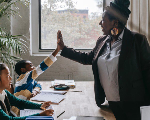 Foto que ilustra matéria sobre melhores escolas em Recife mostra uma professora negra fazendo um gesto de toca aí com um aluno, enquanto uma aluna ao lado observa
