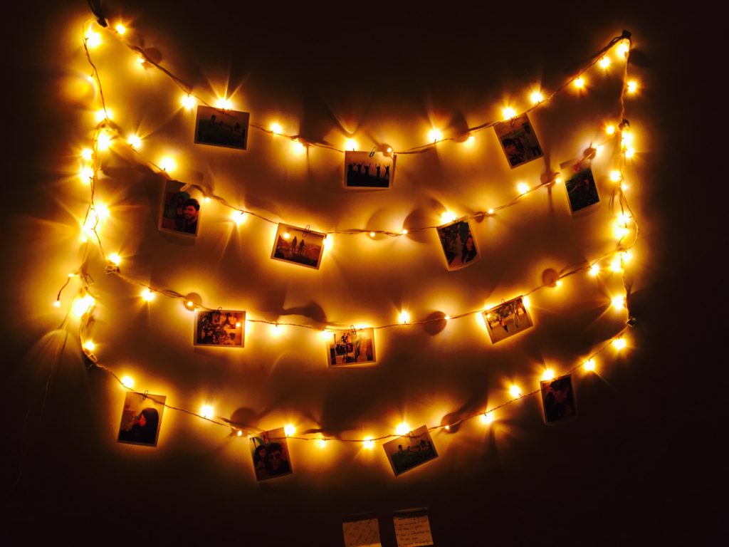 Foto ilustra um espaço escuro com um varal e diversas fotos penduradas. No varal também tem algumas luzinhas. 