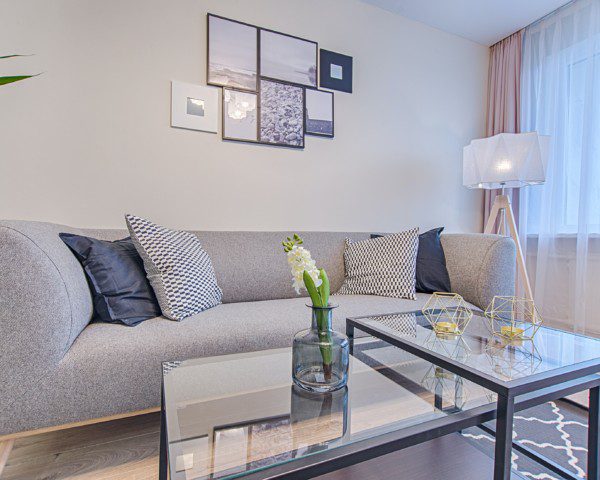 A foto ilustra uma decoração de sala de estar, contendo um sofá cinza, com uma parede bege ao fundo com alguns quadrinhos pendurados na parede. Além disso, conseguimos observar uma mesa de centro de vidro e uma luminária de chão.