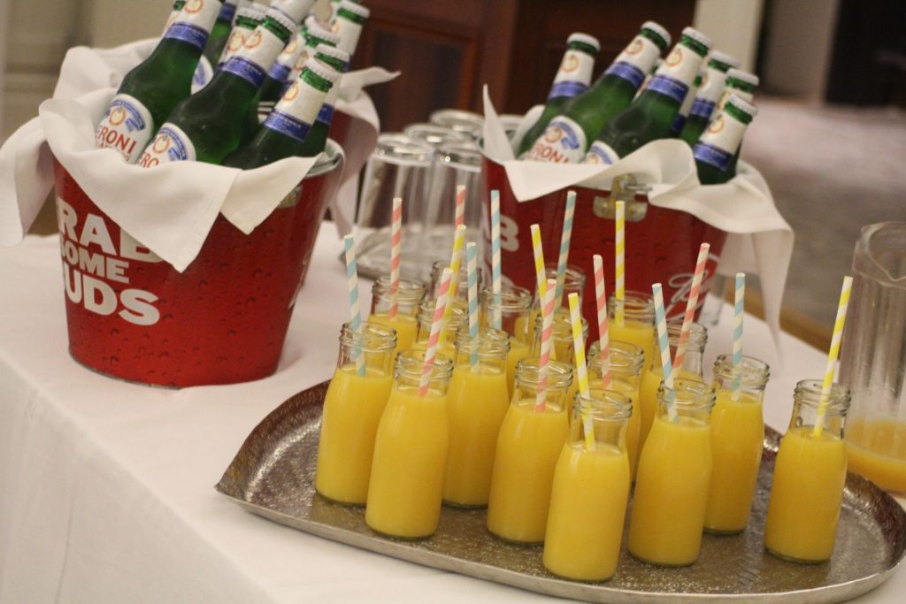 Foto ilustra uma mesa com uma bandeja e garrafas de suco com canudos coloridos e também baldes com cervejas. Ótima opção de barzinho para decoração de chá de panela.