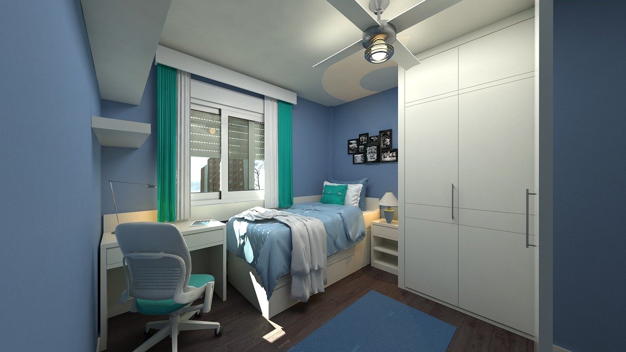 Imagem de um quarto pequeno de um adolescente. Possui uma cama de solteiro, um armário branco com porta de correr, paredes na cor azul e uma mesinha branca no canto esquerdo com um notebook.