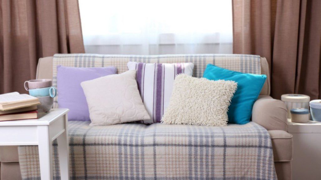 manta centralizada no sofá, com almofadas e, atrás, janela e cortinas