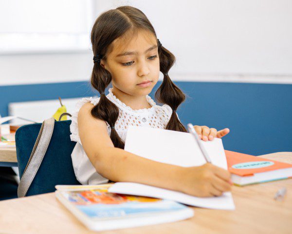 Foto que ilustra matéria sobre Escolas em Fortaleza mostra uma menina sentada com um caderno aberto à sua frente e escrevendo com um lápis em uma sala de aula.