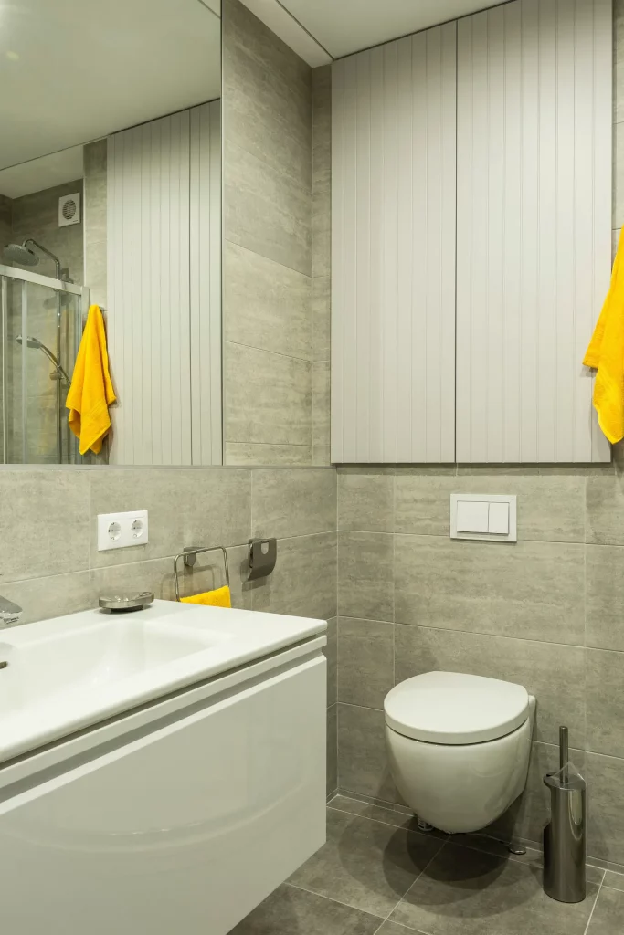  Imagem de um lavabo moderno com um vaso sanitário na cor branca. O lavabo é todo revestido, tem persianas na janela, uma pia com gabinete e um espelho na parede.