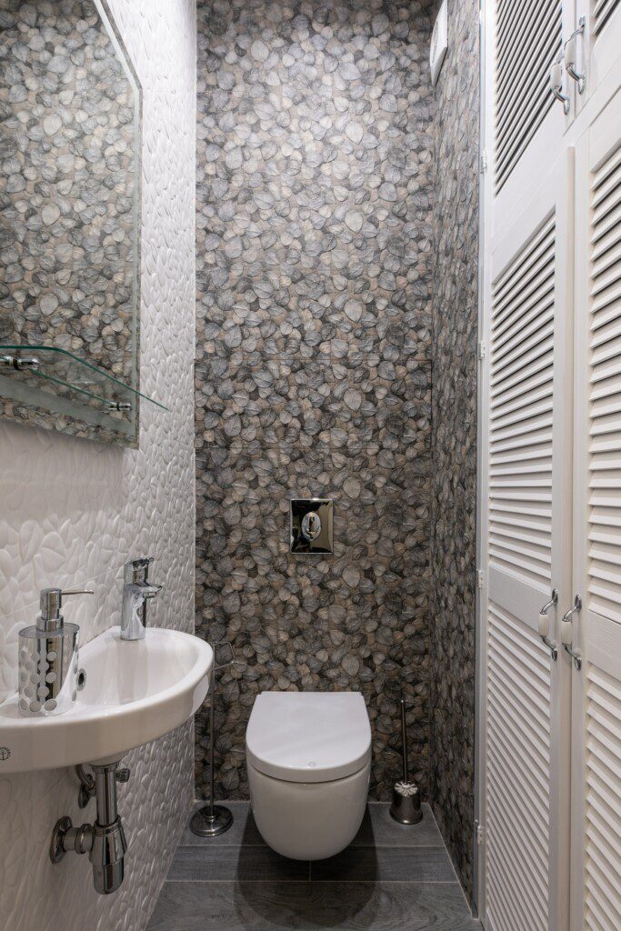 lavabo pequeno com pia, espelho, armário com portas, vaso sanitário e parede feita com pedras. 
