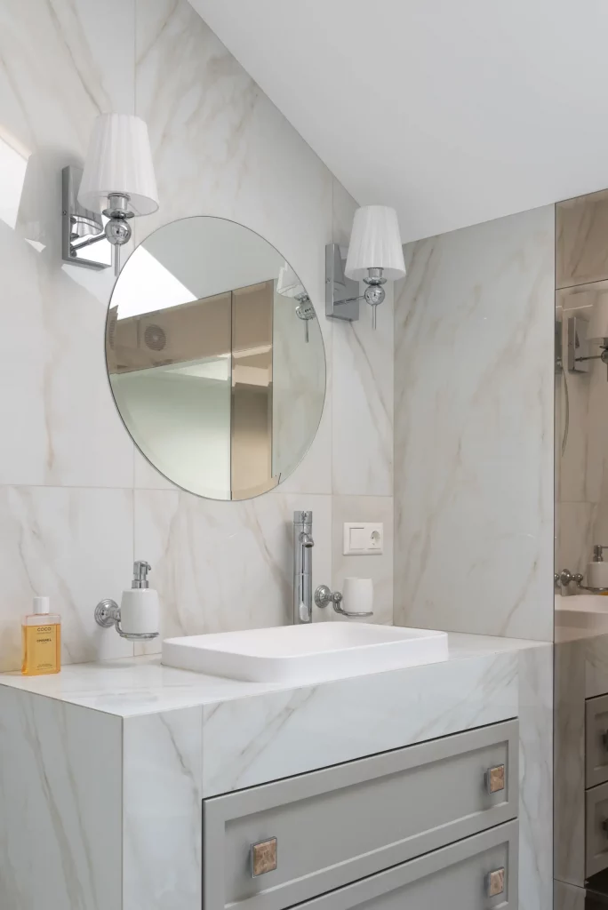 A imagem mostra um lavabo pequeno em tons claros. Nele há uma pia em formato retangular em cima de uma bancada de pedra branca, um espelho oval e dois lustres de parede.