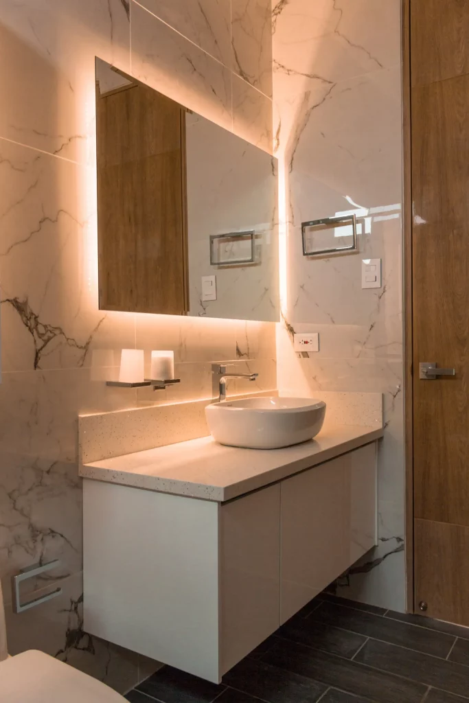 A imagem mostra um lavabo com iluminação especial. Nele há uma pia com armário embutido embaixo, espelho retangular com luz especial ao redor e um suporte para toalha de rosto.
