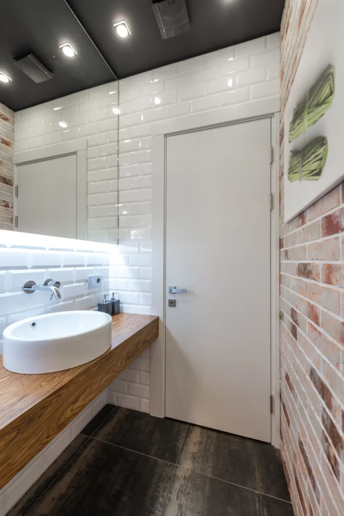 A imagem mostra um lavabo com um papel de parede em uma das paredes. Nele há uma pia em formato oval em cima de uma bancada de madeira rústica, um espelho retangular e um papel de parede que imita tijolinhos.