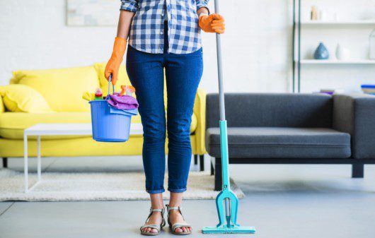 Imagem de uma mulher com calça jeans e uma camisa xadrez azul pronta para limpar sofá de tecido. Ela está com um balde e alguns produtos de casa e no fundo há 2 sofás: um amarelo e outro cinza.