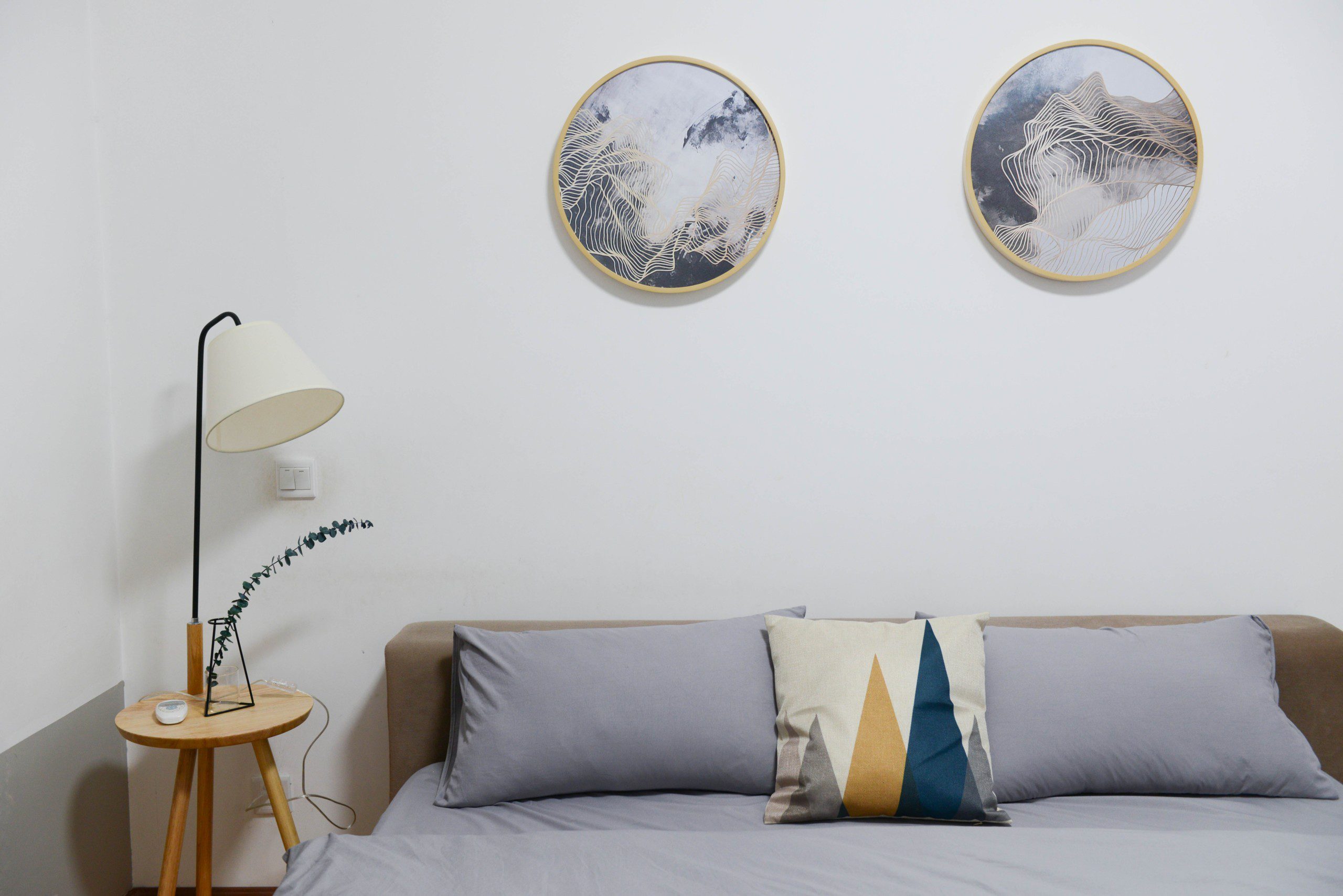 Imagem do canto de um quarto de casal com uma banco que está sendo usado como mesinha de cabeceira. Em cima do banco há um abajur e na parede um quadro com formas geométricas. 